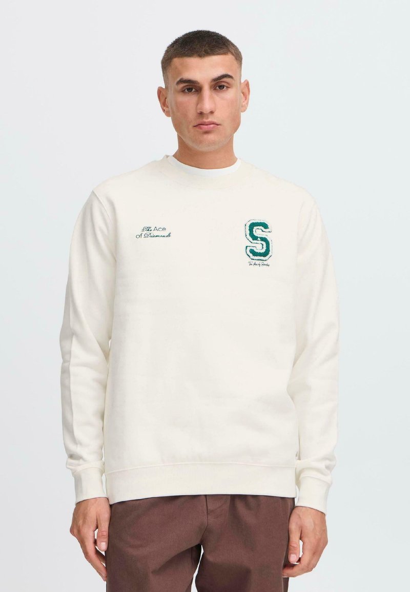 Hanorac crem cu manșete și tiv cu nervuri, având brodat pe piept un „S” verde și text. Modelul poartă pantaloni maro. Design simplu.
