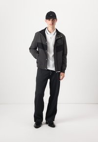 Giacca nera con pannelli scuri, polo bianca, pantaloni neri e berretto nero. L'outfit presenta una vestibilità rilassata e un design minimalista.