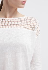 Bílý dlouhý rukáv top s texturovanou krajkovou vsadkou u výstřihu, s volným střihem a jemnou, lehkou tkaninou. Akcenty v podobě průhledných detailů.