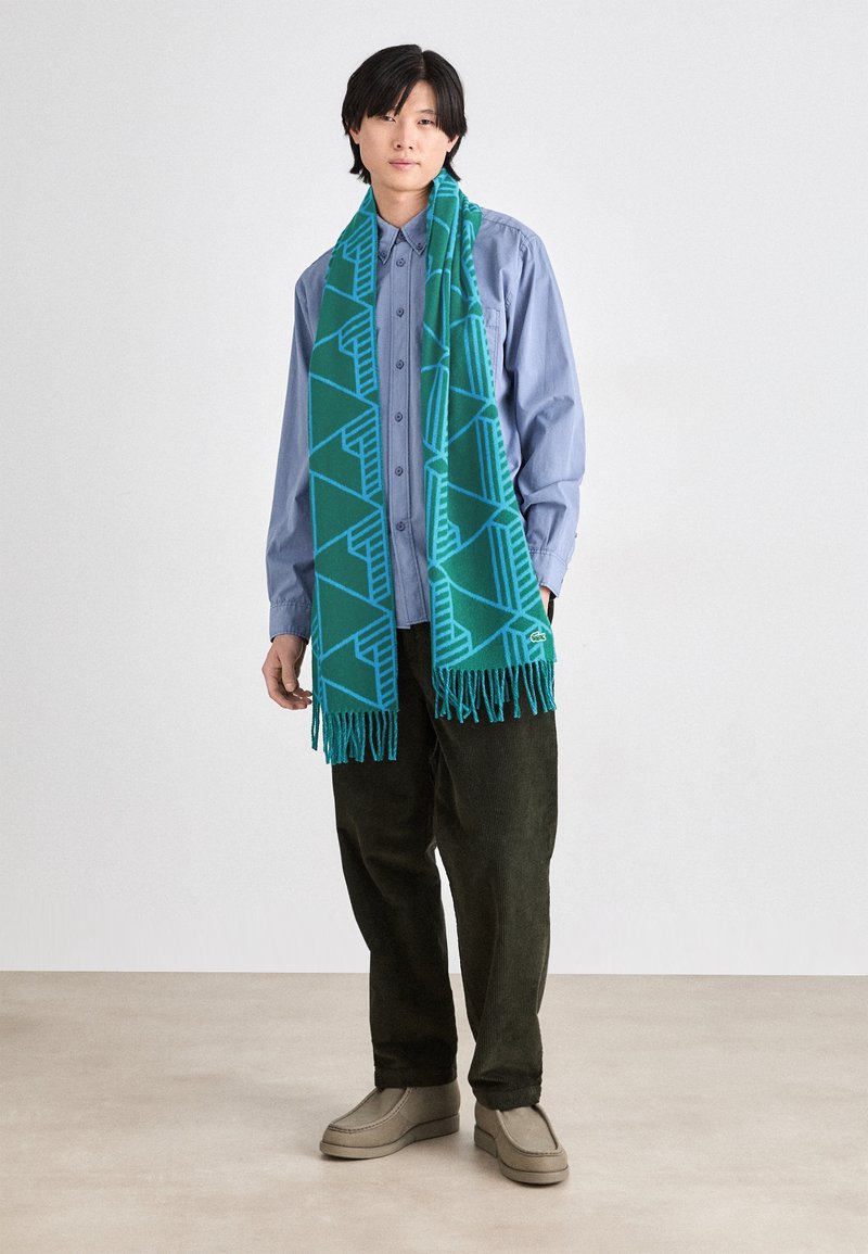 Bufanda verde azulado con un patrón geométrico, con bordes desflecados, hecha de material suave, llevada sobre una camisa de botones azul, con pantalones oscuros y zapatos grises.