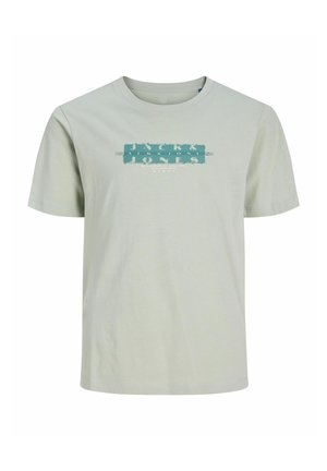 Blond jongetje staat, draagt een lichtgroen grafisch T-shirt, zwarte cargobroek en witte sneakers tegen een egale witte achtergrond.