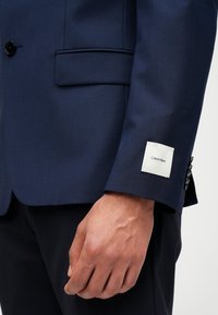 Jūrinis švarkas su glotnia tekstūra, vienu mygtuku, ir baltu etikete su užrašu "Calvin Klein" ant rankovės.