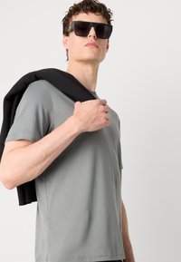 Tricou gri cu mânecă scurtă, cu o textură netedă, asortat cu ochelari de soare negri supradimensionați. Un pulover închis este aruncat peste o umăr.