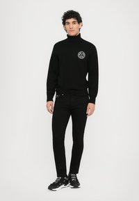 Νεαρός άνδρας φορώντας μαύρο πουλόβερ με τσάκιση, μαύρα στενά τζιν και μαύρα αθλητικά παπούτσια, στέκεται μπροστά σε έναν απλό λευκό τοίχο.