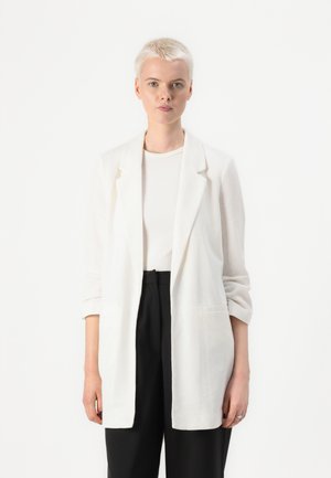 Bílý lehký blazer s volným střihem, klopami a dvěma bočními kapsami. Nositý přes světlo barevný top a kombinovaný s tmavými kalhotami.