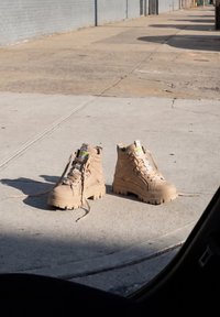 Pair of beige lace-up boots with thick soles placed on a sunlit concrete sidewalk near a building wall.