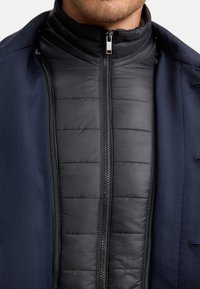 Herenjas met een zwart geïsoleerd binnenlaag met een gequiltde textuur, in contrast met een marineblauwe buitenlaag, beide met ritsen en hoge kraag.