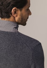 Donkergrijze gebreide trui met ribbelstructuur en een contrasterende grijze kraag, met een gladde afwerking en gestructureerde schoudernaden.