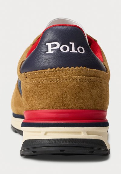 Smeđe antilop tenisice s mornarsko plavim kožnim petama, crvenim akcentima i bijelim "Polo" logom. Imaju podstavljeni đon i teksturirani crni potplat.