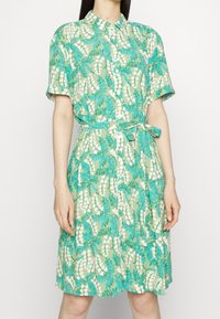 Knielanges, hellgrünes Hemdkleid mit türkis-weißer Blütenmusterung. Kurze Ärmel, tailliert mit Gürtel.