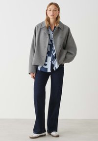 Giacca cropped grigia di lana con maniche ampie, due bottoni, indossata sopra una camicia a righe blu e bianca; abbinata a jeans svasati scuri e sneakers.