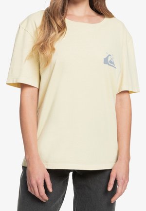 Vrouw draagt een losse lichtgele T-shirt met een klein blauw golflogo op de linkerborst, gecombineerd met donkergrijze broek.
