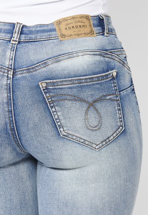 Jeans en denim bleu clair avec une étiquette en cuir beige de marque, des coutures décoratives sur la poche arrière et une silhouette ajustée.