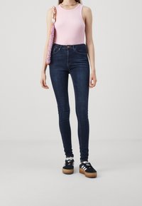 Jeans oscuros de corte ajustado combinados con una blusa sin mangas rosa. La modelo lleva zapatillas negras con suela de plataforma. El conjunto incluye un bolso de punto colorido.