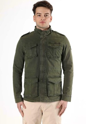 Un homme pose avec une veste utilitaire vert olive à quatre poches à rabat et un pantalon beige sur un fond blanc uni.