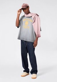 Grijze oversized T-shirt met 'INDIANA STATE' in geel, roze sweatshirt over de schouder gehangen, donkerblauwe spijkerbroek en tankleurige sneakers.