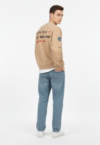 Felpa beige con design ricamati, inclusi testo e accenti colorati, abbinata a jeans azzurri chiari e sneakers bianche.