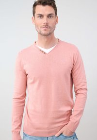 Pull en V rose clair à manches longues, avec trois boutons décoratifs au niveau du col et un ourlet côtelé.