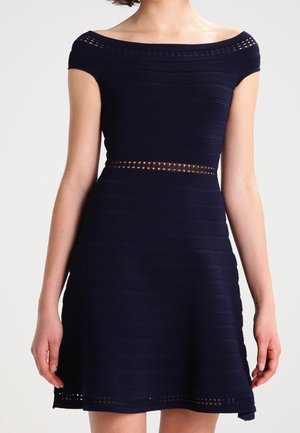 Vrouw draagt een marineblauwe, off-shoulder, knielange jurk met horizontale ribbels en perforaties bij de taille, halslijn en zoom.