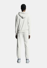 Haut à capuche blanc avec cordon de serrage, jogging assorti, texture douce, coupe décontractée, poignets côtelés, porté avec des chaussures de sport grises.