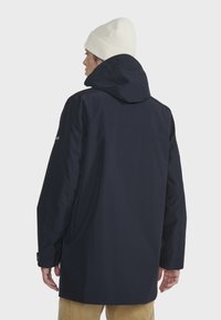 Marineblauwe waterdichte jas met een capuchon, zijzakken en elastische manchetten, gecombineerd met een witte gebreide muts. Soepele textuur, minimalistisch ontwerp.