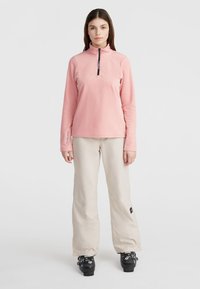 Pink fleece half-zip top with long sleeves, featuring a zippered collar and branding on the sleeve; paired with beige snow pants and black boots.