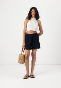 Strikket hvit crop top og marineblå shorts med blonder, holder en vevd stråveske. Sorte flip flops fullfører det uformelle antrekket.