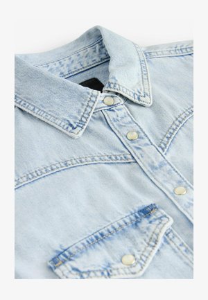 Hele sinine teksapood, millel on klassikaline krae, metallist nupud, tasku detail ja tekstureeritud kangas. Omab kontrastset õmbluse disaini.