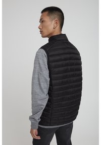 Schwarze Daunenweste mit horizontalem Steppmuster, hohem Kragen und ohne Ärmel, über einem grauen Langarmshirt getragen. Glatte Textur.