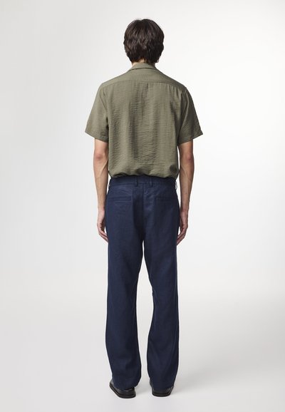 Camicia verde oliva a maniche corte con una texture sottile, abbinata a pantaloni blu navy dal taglio morbido e scarpe nere. Vista posteriore mostrata.
