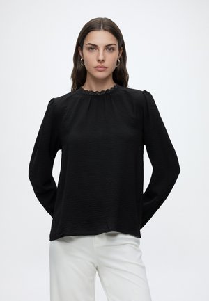 Femme aux longs cheveux bruns portant un chemisier noir à manches longues avec un col en dentelle et un pantalon blanc, faisant face avec les mains dans le dos.