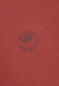 Ύφασμα χρώματος σκουριάς με κεντρικό κυκλικό λογότυπο που περιλαμβάνει ένα δέντρο και κείμενο "BLKVIS EST. 2013" σε σκούρο τύπωμα.