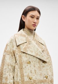 Jauna sieviete ar taisniem tumšiem matiem, valkājot bēša toņa dubultpogas mēteli pār gaišu augstu apkakli, uz vienkrāsaina fona.