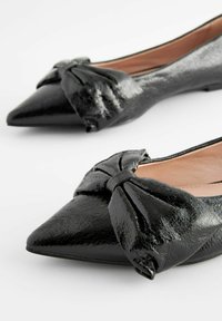 Musta patenttinaapuri, jossa on terävä kärki ja nauhadesign etuosassa. Silkinsmooth pinta kiiltävällä pinnalla. Yksinkertainen, elegantti muotoilu.
