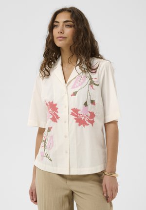 Vrouw met krullend bruin haar, gekleed in een wit overhemdbloesje met korte mouwen met roze en rode bloemenpatronen en beige broek.