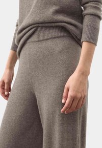 Primo piano di una persona che indossa un maglione a maglia taupe e pantaloni larghi coordinati sempre a maglia, con cintura e polsini a costine.