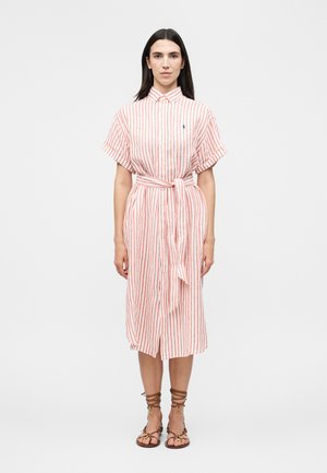 Femme portant une robe blanche mi-longue à rayures verticales rouges, manches courtes, ceinture nouée devant et sandales marron à lacets, debout, droite.