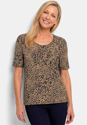 Femme souriante aux longs cheveux blonds portant un haut à manches courtes imprimé animal marron et noir et un pantalon foncé sur fond clair.