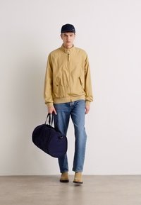 Lys beige bomberjakke, blå jeans, tan støvler og en navy duffel taske. Skarpt design med minimalt detaljer, stående foran en ensfarvet baggrund.