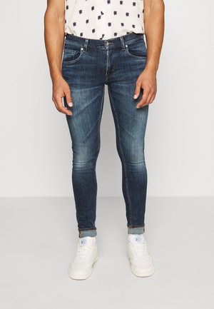 Homme portant un jean slim bleu foncé avec les revers roulés, des chaussettes blanches et des baskets blanches, debout devant un fond clair uni.