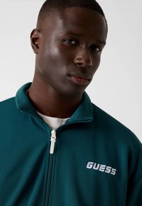 Petrolblauwe jas met ritssluiting en een getextureerd oppervlak, met een wit "GUESS"-logo op de linkerborst en een witte ritstrekkertrekker.