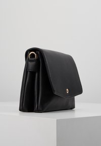 Černá kožená křížová taška s detailem zlatého kroužku, obdélníkovou klopou a jediným zapínáním na knoflík, umístěná na bílé ploše s čistým pozadím.