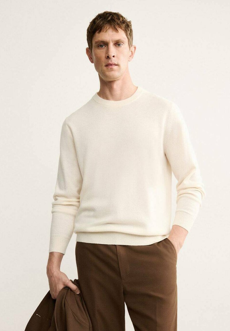 Maglione color crema con collo rotondo e maniche lunghe, realizzato in un materiale morbido. Abbinato a pantaloni marroni, completato da una giacca casual.
