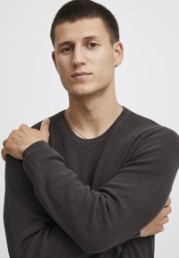 Ciemnoszary sweter dziany o ribbowanej fakturze, z okrągłym dekoltem i dopasowanymi rękawami. Modelka pozuje z założonymi rękami, prezentując srebrny naszyjnik.