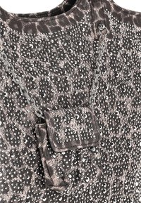 Metallinhohtoinen harmaa kangas, jossa on teksturoitu kuvio ja hajallaan hopeisia strasseja. Pienessä kiinnitettyssä taskussa on ketjuhihna.