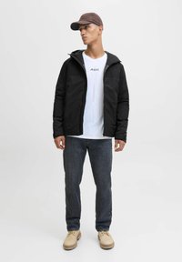 Czarna kurtka z zamkiem, biała koszulka z logo, ciemne dżinsy i beżowe buty na sznurówki. Brązowa czapka dopełnia outfit. Neutralne tło.