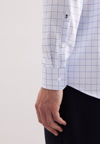 Camicia bianca con motivo a reticolo blu, polsini abbottonati, design ricamato sulla manica, tessuto di cotone testurizzato, primo piano di una mano che tiene la camicia.