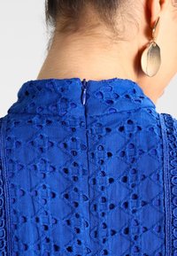 Tela azul bordada con patrones de cortes, presenta una cremallera trasera y detalles texturizados cerca del escote. Pendiente grande de oro visible.