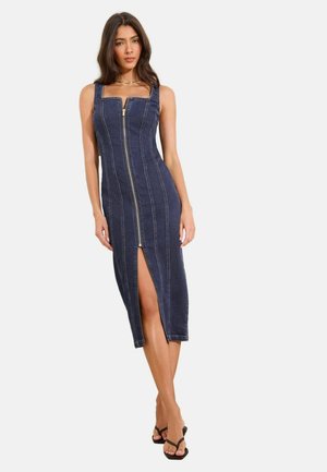 Femeie purtând o rochie midi mulată din denim închis la culoare, fără mâneci, cu fermoar frontal și fustă cu despicătură în față, asortată cu sandale negre cu barete.