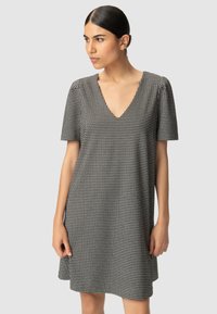 Kobieta z ciemnymi włosami w niskim kokie, ubrana w czarno-białą sukienkę w wzór hounds-tooth z dekoltem w serek i krótkim rękawem, patrząca w bok na jasnoszarym tle.